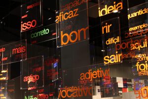 Foto de uma sala da exposição Menas com palavras diversas penduradas em painéis acrílicos transparentes.