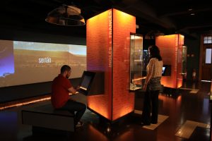 Foto de uma sala do Museu da Língua Portuguesa onde dois visitantes observam um computador e uma vitrine.
