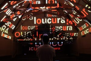 Foto de um visitante observando o teto do museu com projeção da palavra loucura várias vezes..