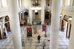 magem do saguão da Estação da Luz a partir do mezanino. Em primeiro plano se vem duas grandes colunas cortando a imagem. No térreo, pessoas circulam pelo espaço, sobre o piso xadrez (branco e cinza claro), com um grande ornamento no meio - um quadrado, com um círculo todo decorado com uma rosácea amarela. Sobre esse elemento, no teto, há um grande lustre pendurado. Nas laterais, no mezanino, um gradil de ferro ornamentado cerca todo o mezanino.