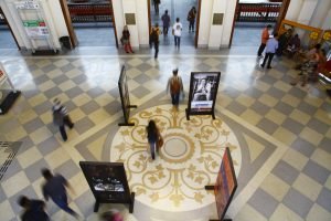 Imagem feita a partir do mezanino da Estação da Luz, mostrando o piso decorado. Pessoas circulam pelo espaço, com piso xadrez (branco e cinza claro) e uma grande rosácea amarela no centro. Há alguns paineis com fotografias espalhados pelo espaço.